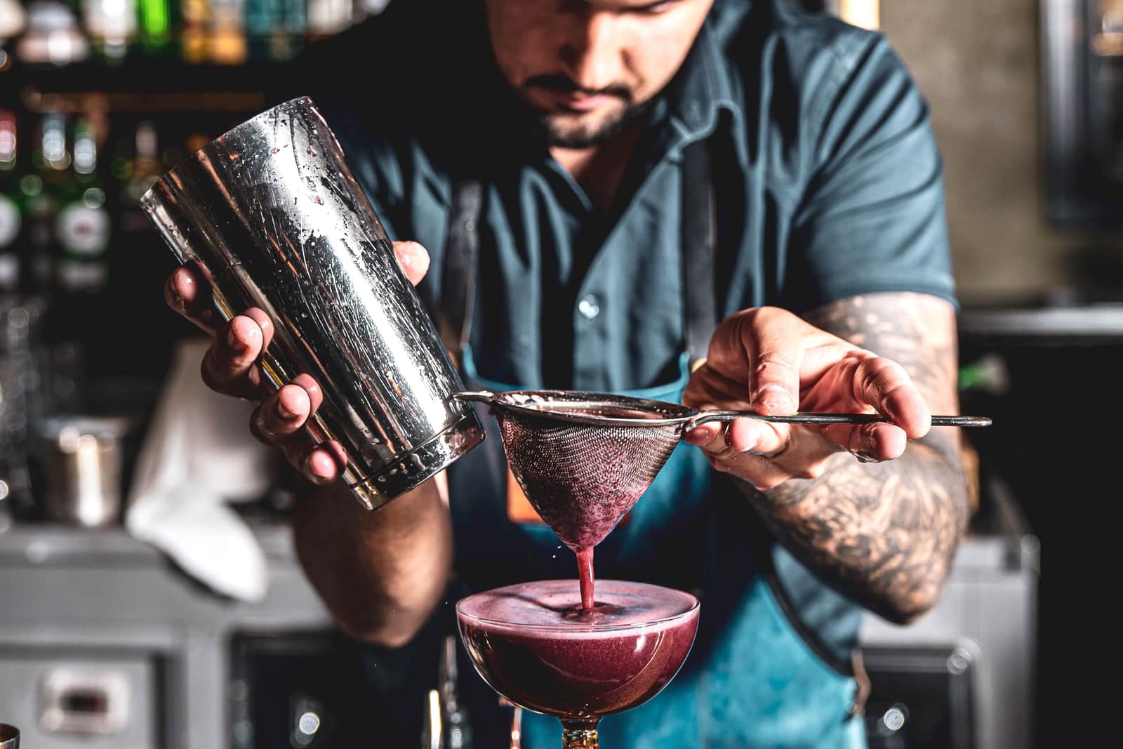 A bartender straining a cocktail with a shaker and strainer on a bar